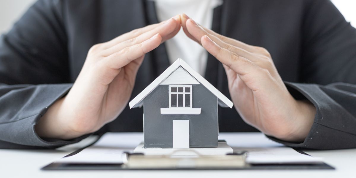 Hands forming a protective gesture over a small house model on a table.