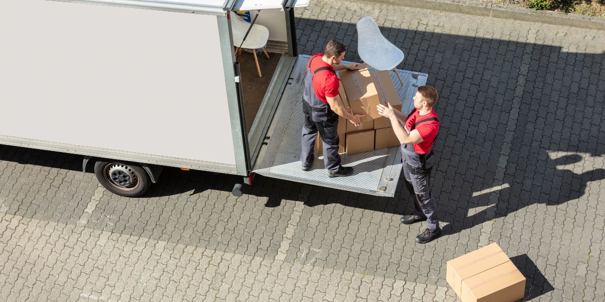 Two movers in red shirts load cardboard boxes onto a moving truck from a liftgate.