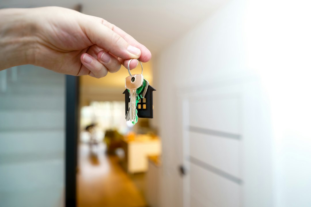 A real estate agent's hand giving a set of modern house keys to a new homeowner, with the beautiful entryway of a luxury home blurred in the background.